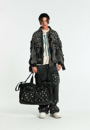 Persona joven con cabello trenzado que lleva chaqueta y pantalones de denim negro con lunares blancos, sosteniendo un bolso grande a juego, zapatillas blancas.