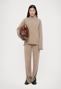 Beige gebreide trui met hoge hals, gecombineerd met bijpassende brede broek. Model houdt een bruine leren tas vast met een gouden hardware detail.