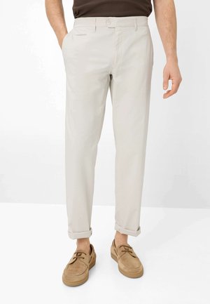 Man die lichtbeige casual broek draagt met opgerolde pijpen en lichtbruine suède veterschoenen, staand tegen een witte achtergrond.