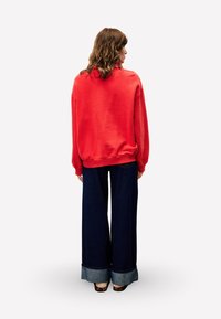 Sweatshirt rouge surdimensionné avec des poignets et un ourlet côtelés, associé à un jean large bleu avec des ourlets roulés et une texture en denim.