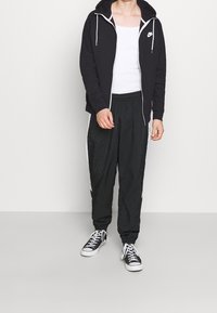 Černá mikina na zip s bílými tkaničkami, doplněná černými tepláky s elastickým pasem, které mají bílé pruhy po stranách, a černými teniskami.