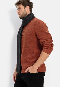 Orange gestrickter Pullover mit einem gerippten Muster, ¼ Reißverschluss am Kragen, kombiniert mit einem dunkelgrauen gerippten Schal, getragen mit schwarzen Jeans.