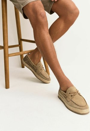 Person, die beige Wildleder-Bootsschuhe und khakifarbene Shorts trägt und auf einem Holzschemel vor weißem Hintergrund sitzt.