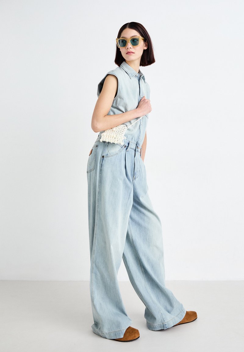 Hellblauer Denim-Jumpsuit mit Kragen und ärmellosem Design, weiten Beinen und einer strukturierten cremefarbenen Tasche. Braune Slipper runden den Look ab.