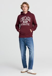 Felpa bordeaux con stampa "CHICAGO 1983 ATHLETIC". Caratterizzata da una tasca a marsupio, cappuccio con stringa, indossata con jeans blu e sneakers bianche.