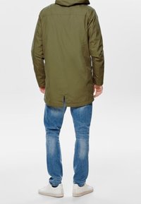 Man i en olivgrön hooded jacka, blå jeans och vita sneakers, som står vänd bort från kameran mot en ljus bakgrund.