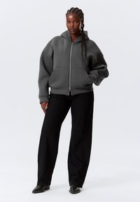 Sweat à capuche gris zippé en tissu doux, offrant une coupe décontractée et de grandes poches avant. Associé à un pantalon noir ample et des bottines.