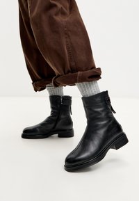 Botas de cuero negras hasta el tobillo con cremallera lateral y detalle de hebilla, llevadas con calcetines de canalé gris y pantalones marrones remangados sobre un suelo claro.