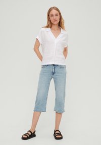 Biała lniana koszula z dekoltem w serek i krótkimi rękawami, w zestawieniu z jasnoniebieskimi, skrojonymi dżinsami. Modelka w czarnych sandałach na ramiączkach.