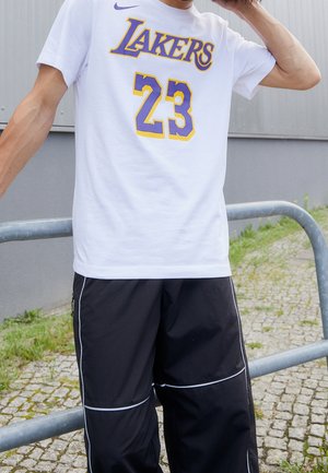 Persona que lleva una camiseta blanca de los Lakers con el número 23 y pantalones negros, de pie al aire libre cerca de una barandilla de metal y un suelo pavimentado.