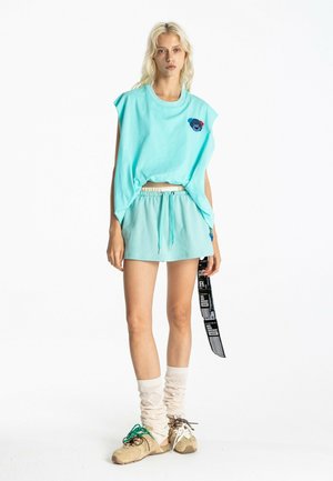 Joven mujer con top sin mangas turquesa con parche de oso, pantalones cortos a juego, calcetines beige arrugados y zapatillas beige sosteniendo una correa en blanco y negro.