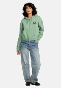 Un hoodie zippé vert menthe en matière douce, avec un design brodé. Associé à un jean bleu clair ample et des baskets noires.