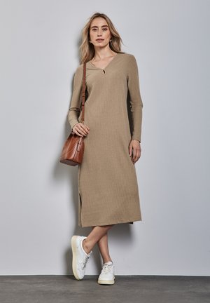 Rippent, langärmliges, beiges Kleid mit V-Ausschnitt und einem seitlichen Schlitz, kombiniert mit weißen Sneakern und einer braunen Umhängetasche.