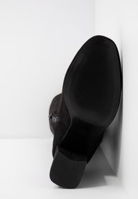 Bottine noire en daim avec une semelle texturée et un petit talon bloc, positionnée avec la semelle vers l'avant sur un fond blanc en coin.