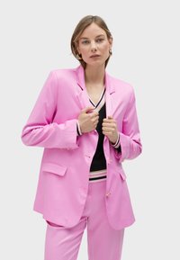 Pink blazer with a structured shape, single-button closure, and two front pockets; paired with a black top featuring striped accents.