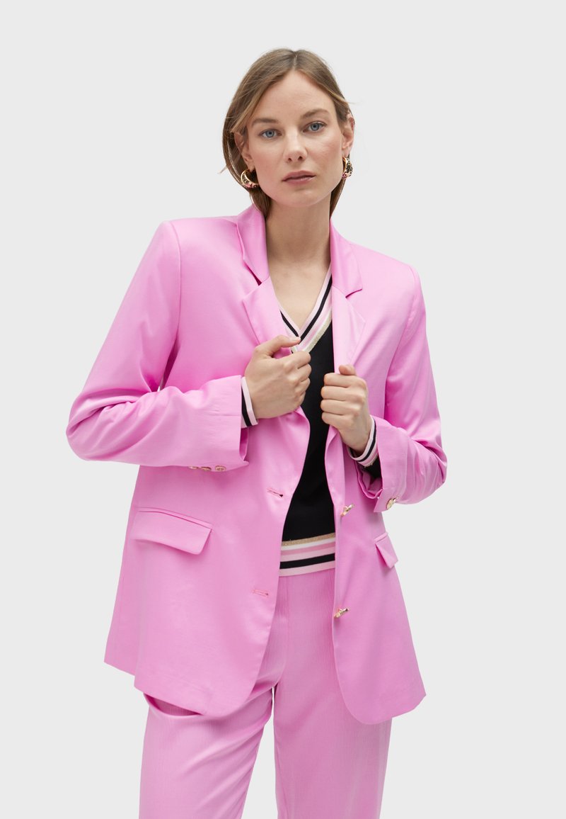 Pink blazer with a structured shape, single-button closure, and two front pockets; paired with a black top featuring striped accents.