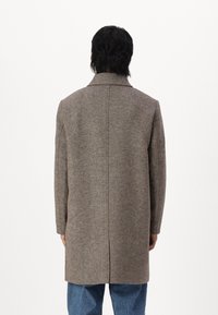 Cappotto marrone, strutturato, con taglio sartoriale, maniche lunghe e spacco posteriore, abbinato a jeans blu. Il tessuto presenta un sottile motivo a trama.