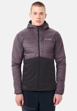 Jeune homme portant une veste matelassée à capuche bicolore violet et noir avec une fermeture éclair à l'avant et une poche sur la poitrine.