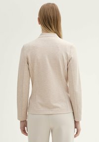 Blazer beige clair en tissu texturé, avec des manches longues, un col mandarin et des détails de couture subtils dans le dos.