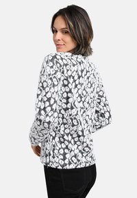 Frau, die sich umdreht, trägt einen hellen und grauen Leopardenprint-Pullover und schwarze Hosen, vor einem schlichten hellen Hintergrund.