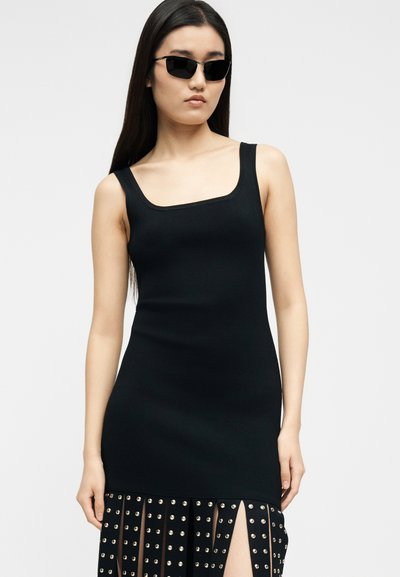 Γυναίκα που φοράει μαύρο αμάνικο φόρεμα με λεπτομέρειες από μεταλλικά τρουκς στη φούστα και μαύρα ορθογώνια γυαλιά ηλίου, στέκεται μπροστά σε απλό γκρι φόντο.