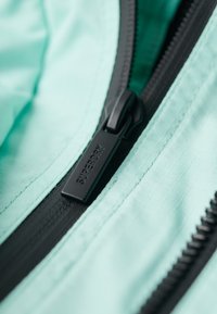 Close-up af en sort Superdry lynlåstrækker på et lyseblåt/turkis stofstykke med en sort lynlåsskinne.