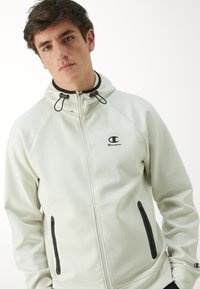 Hellgraue Zip-Hoodie aus weichem Material, mit schwarzem Logo und Reißverschluss-Details, ausgestattet mit verstellbaren Kordeln an der Kapuze.