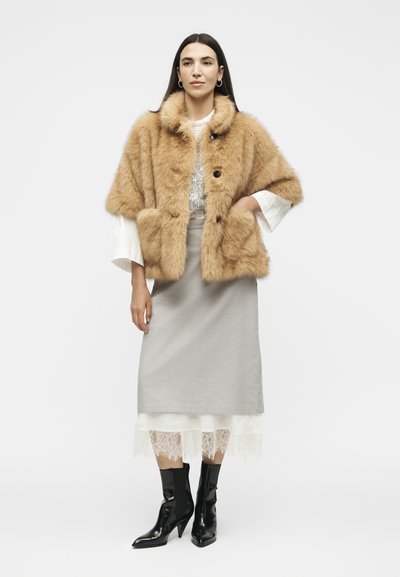 Femme portant un manteau en fourrure beige par-dessus un chemisier blanc avec une jupe grise, un bord en dentelle et des bottines à talons noires, debout devant un fond uni.