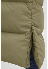 Veste rembourrée vert olive avec fentes sur les côtés, fermetures à pression et surface texturée. Le matériau est léger et doux.