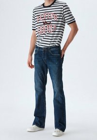 Conjunto de hombres que incluye una camiseta de rayas blancas y negras con letras rojas y jeans azules. Las zapatillas son blancas. El denim muestra un ligero desgaste.