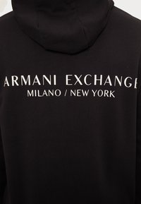 Sweat à capuche noir avec l'inscription "ARMANI EXCHANGE MILANO / NEW YORK" en grand texte blanc. Tissu doux avec une coupe standard et une poche kangourou.