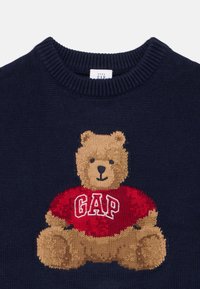 Marinblå stickad tröja med en broderad tan-färgad björn som bär en röd skjorta med texten "GAP". Rundad halsringning och träig textur.