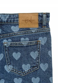 Džinsai su šviesiai mėlyna širdelių raštu, ant jų yra rudos odos lopas su užrašu „VERO MODA“ ir nugarinėje kišenėje matoma siūlė.