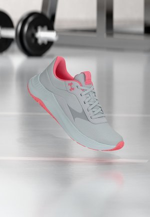 Lichtgrijze en roze sportschoen met veters, merklogo en getextureerde zool, afgebeeld alsof hij zweeft in een sportschool met wazige gewichten op de achtergrond.