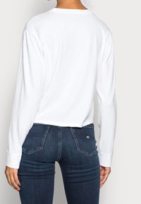 Vit långärmad skjorta med rundad halsringning, cropped design, i kombination med mörkblå tajta jeans med bakfickor och lätt slitningar.