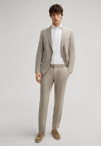 Abito beige su misura con giacca aderente, pantaloni slim e camicia bianca. Tessuto strutturato e lucentezza sottile, abbinato a mocassini marrone chiaro.