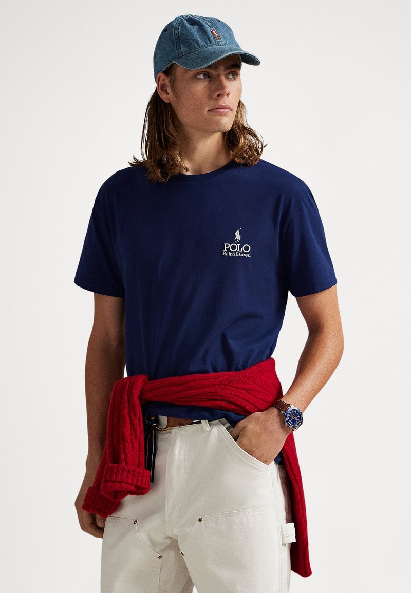 Μπλε βαμβακερό t-shirt με λευκό λογότυπο "Polo Ralph Lauren", συνδυασμένο με κόκκινο πλεκτό πουλόβερ πλεξούδα δεμένο γύρω από τη μέση και λευκό παντελόνι.