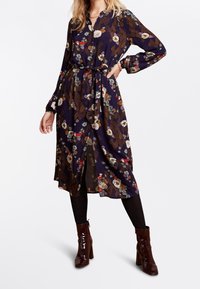 Blommig midiklänning i marinblått medknappstängning framtill, långa ärmar, knytmidja och med en mix av blommor och geometriska mönster. Bärs med svarta strumpbyxor och bruna ankelskor.