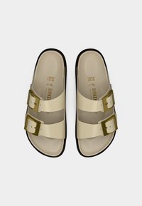 Δύο μπεζ σανδάλια τύπου σλάιντ με δύο ρυθμιζόμενες αγκράφες, λεία υφή και επενδυμένο πέλμα, τοποθετημένα σε καφέ εξωτερική σόλα. Μέγεθος 39.