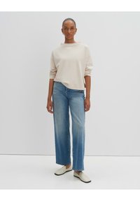 Felpa oversize beige, jeans a gamba larga blu chiaro e scarpe slip-on bianche, su uno sfondo neutro.