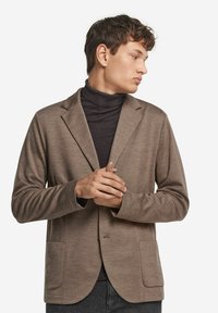 En ung man som bär en brun kavaj över en svart polotröja, tittar åt höger med händerna sammanfognade framför en vit bakgrund.