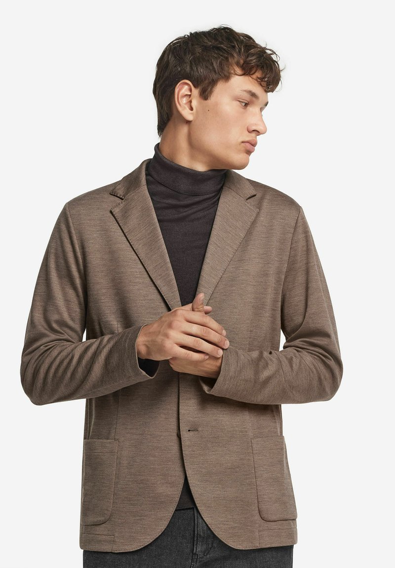 En ung man som bär en brun kavaj över en svart polotröja, tittar åt höger med händerna sammanfognade framför en vit bakgrund.