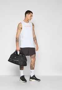 White sleeveless athletic tank top with red "HYROX" logo, black shorts, black duffel bag, and dark sneakers with green accents.