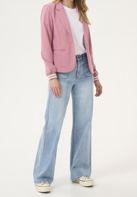 Blazer rose avec coupe structurée, bouton unique et deux poches avant. Associé à un jean large bleu clair et des baskets blanches.