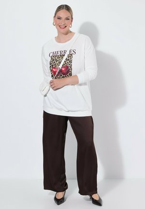 Donna sorridente in piedi, con felpa bianca con stampa di ciliegie, pantaloni larghi scuri, tacchi neri, orecchini a cerchio dorati e piccola borsa bianca a tracolla.