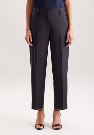 Femme portant un pantalon droit noir sur mesure et des talons noirs à bride cheville et bout ouvert, debout devant un fond uni.