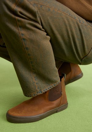 Person trägt braune Leder-Stiefeletten und grünlich-braune Hosen und hockt auf einer hellgrünen Oberfläche.