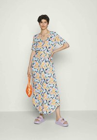 Jovem com um vestido midi floral de mangas curtas, segurando uma bolsa de rede laranja, usando sandálias de plataforma roxas, em pé contra um fundo simples.