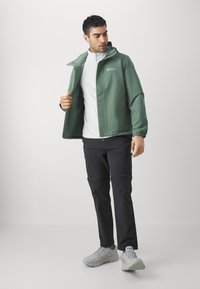 Veste imperméable verte avec fermeture éclair à l'avant et poignets élastiques, portée sur un sweat-shirt blanc et un pantalon cargo noir. Chaussures gris clair.