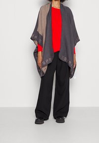 Poncho en laine tricotée grise et beige avec des accents de logo, au design ouvert, présentant une texture douce et une silhouette drapée, porté sur un haut rouge.